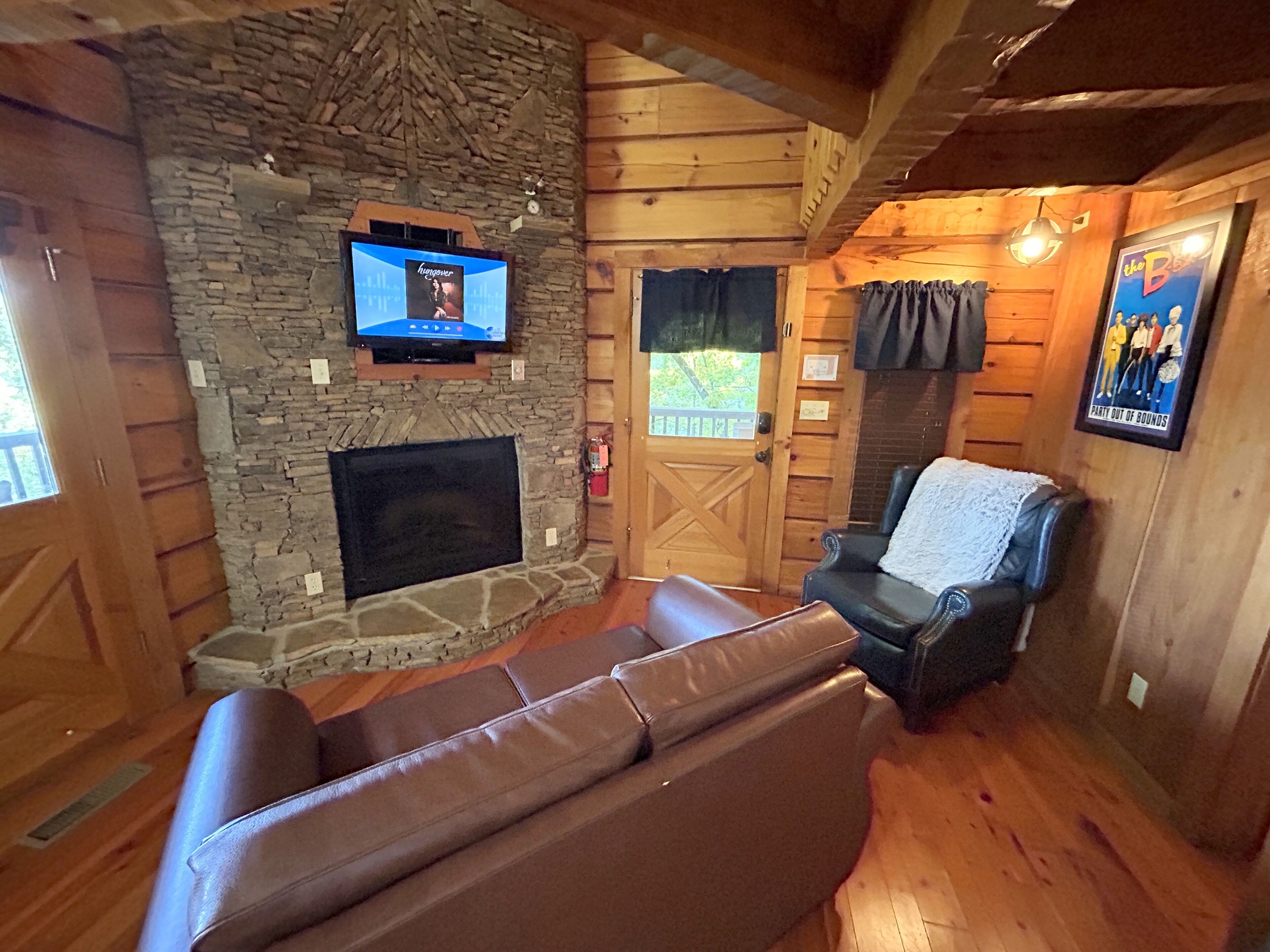 Ein gemütliches Blockhaus-Interieur mit Kamin aus Stein, einem darüber montierten Fernseher, einer Ledersofa und einem gerahmten Poster an der Wand.