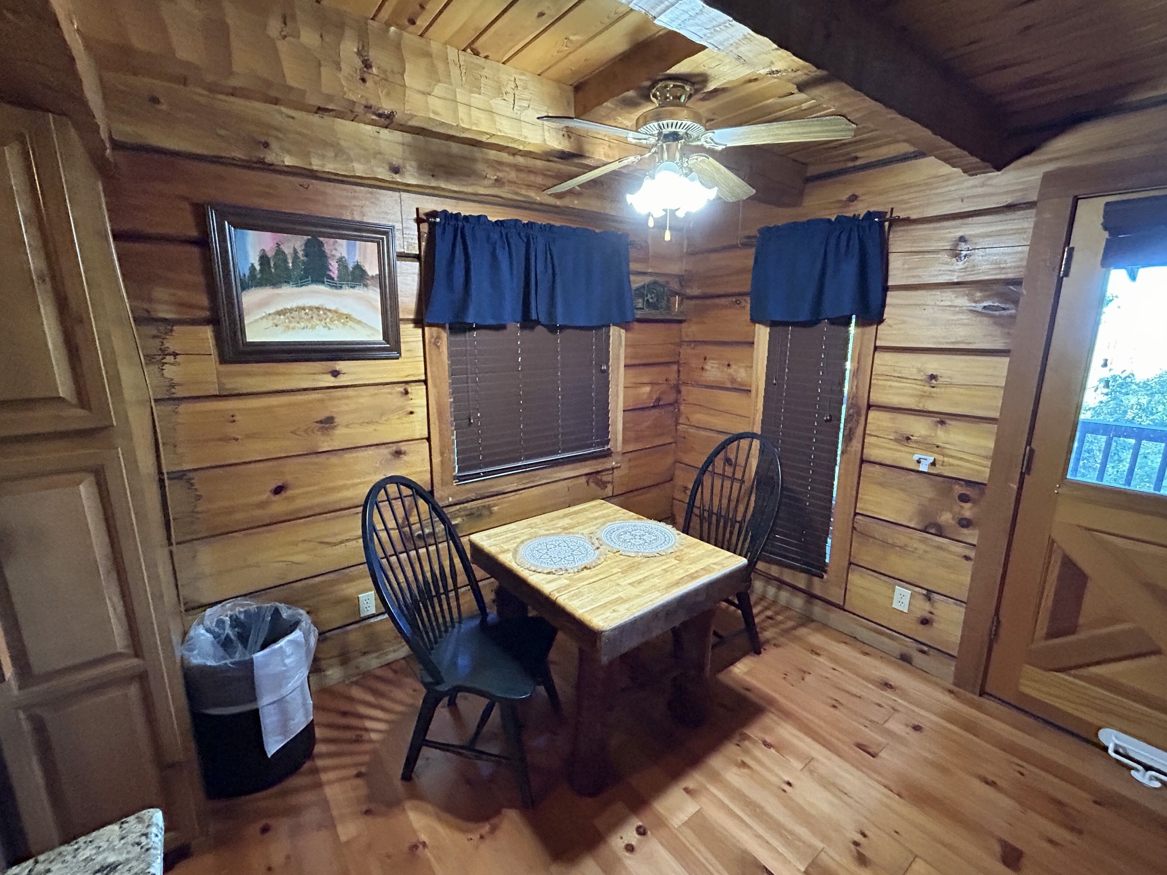 A wooden cabin interior with a dining table, two chairs, a ceiling fan, and a picture on the wall.