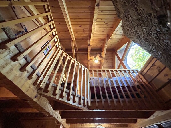 A wooden spiral staircase in a cabin with a ceiling fan, a window with a view of greenery, and a stone wall. - Love Shack honeymoon cabin that is stairway to the stars! (Sevierville)