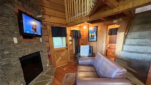 Interior acogedor de una cabaña con chimenea de piedra, sofá de cuero, paredes de madera y televisor montado.