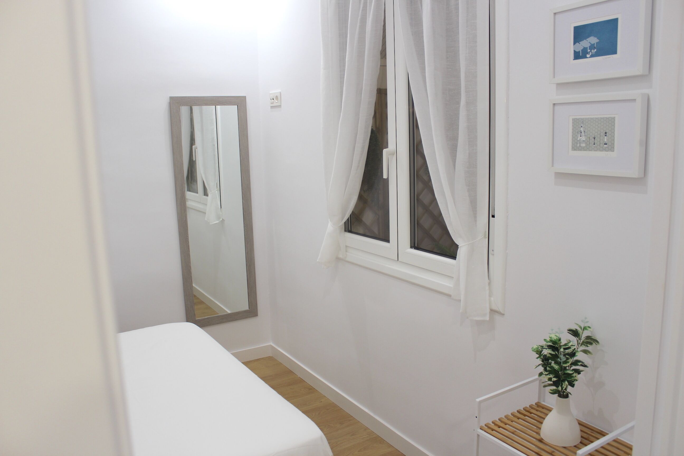 A small, clean room with a single bed, a window with curtains, a mirror, and a small plant on a shelf.
