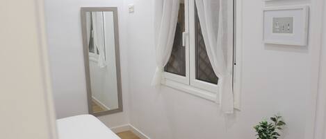 A small, clean room with a single bed, a window with curtains, a mirror, and a small plant on a shelf.