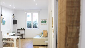 Ein modernes Wohnzimmer mit einer Couch, einem Couchtisch und einem Fernseher auf einem Schrank. Ein Fenster bietet Blick auf eine grüne Umgebung.