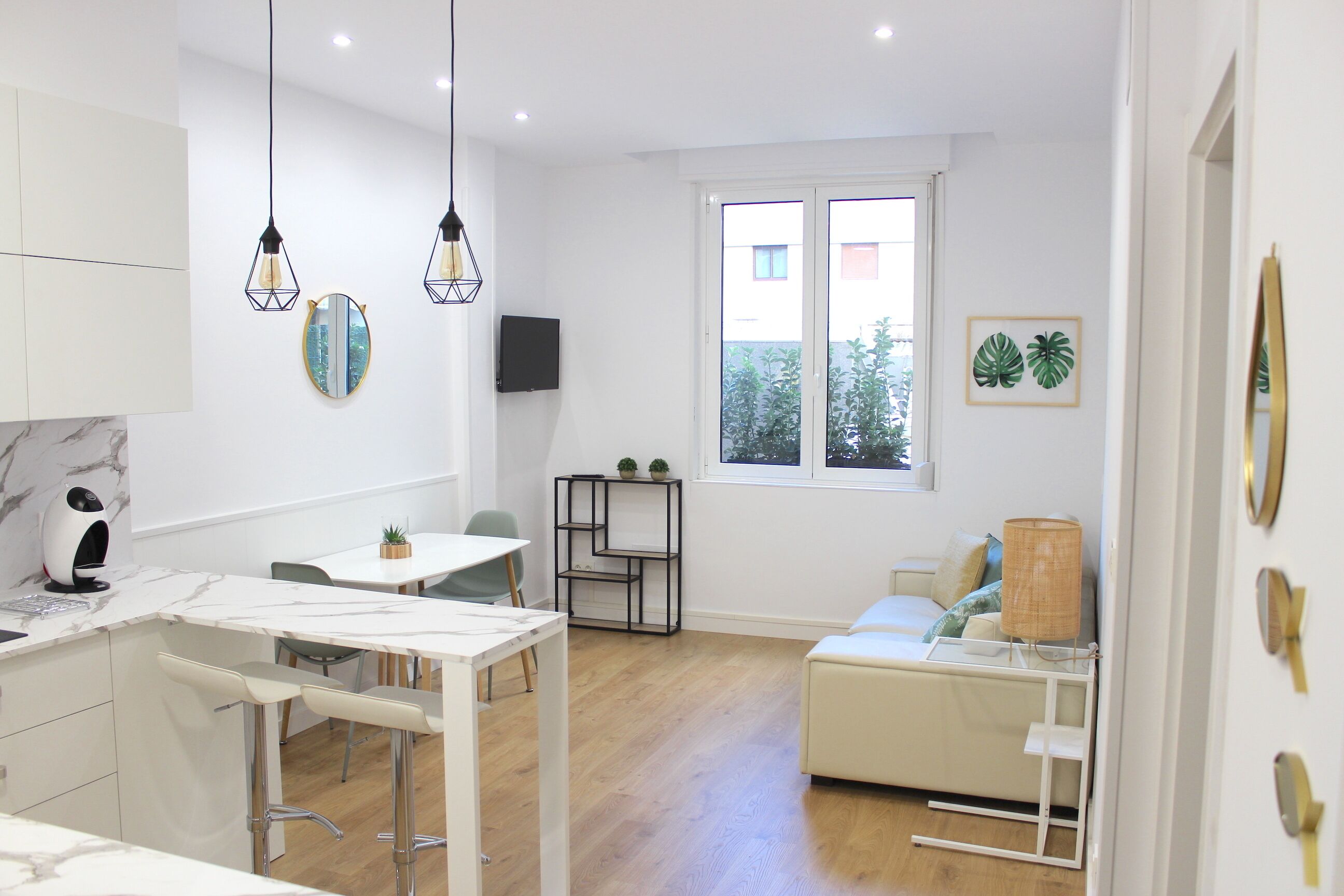 A modern kitchen with white cabinetry, a marble countertop, and pendant lighting. There is a dining area with a white table and chairs, and a small seating area with a white sofa and a lamp.