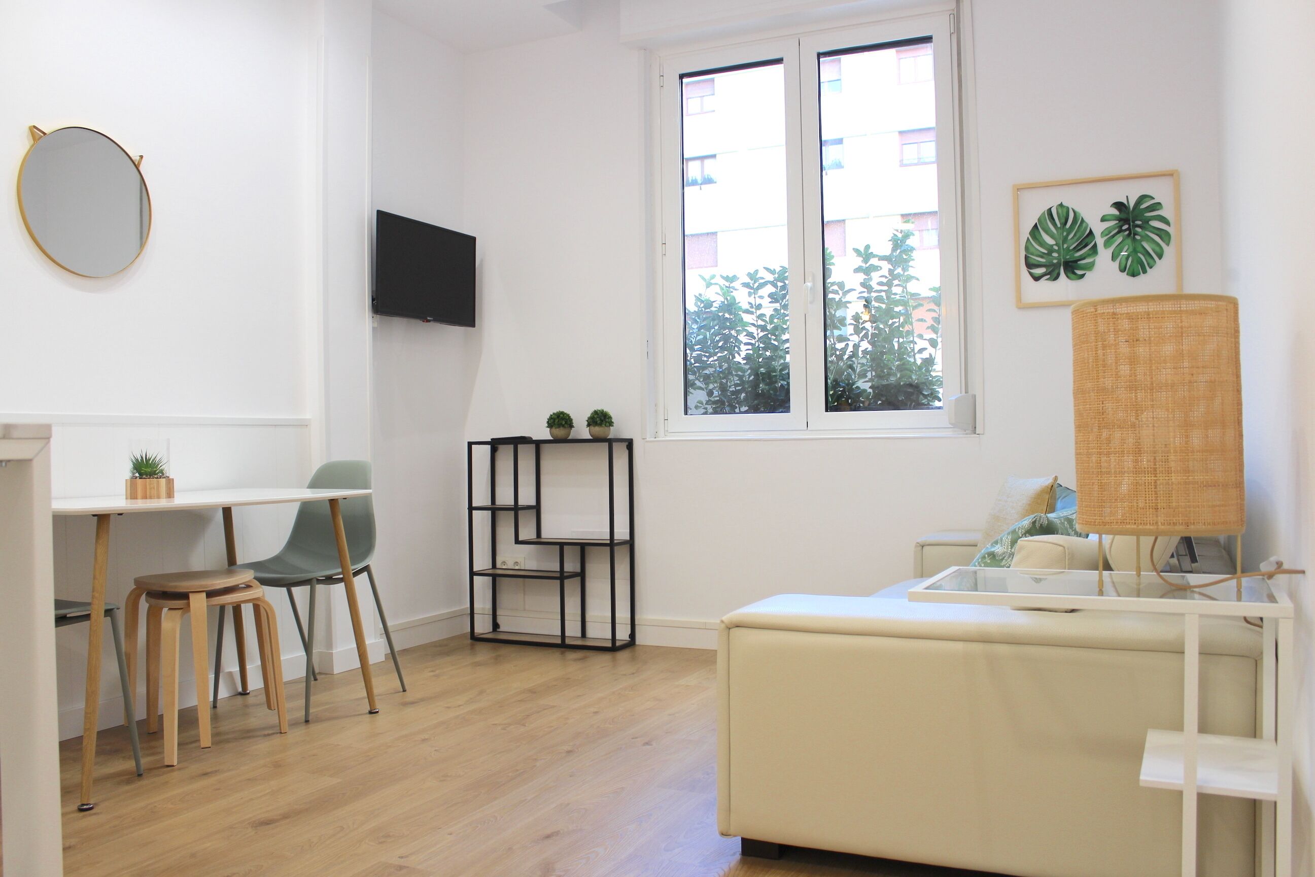 A room with a desk, two chairs, a small table, a sofa, a TV, a window, and two potted plants.