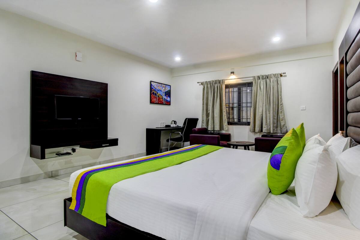 A hotel room with a large bed, a flat-screen TV mounted on the wall, a desk with a chair, and a window with curtains.