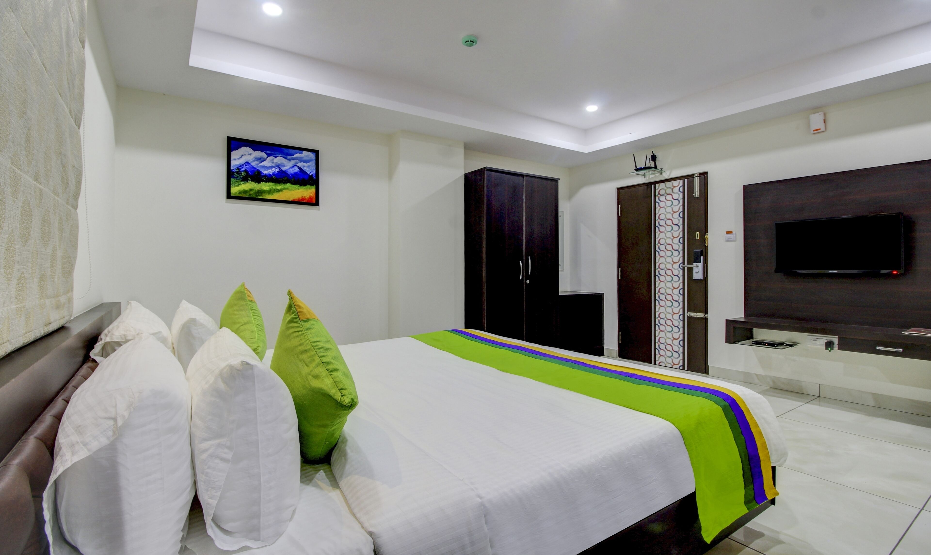 A hotel room with a bed, pillows, a TV, a wardrobe, and a painting on the wall.