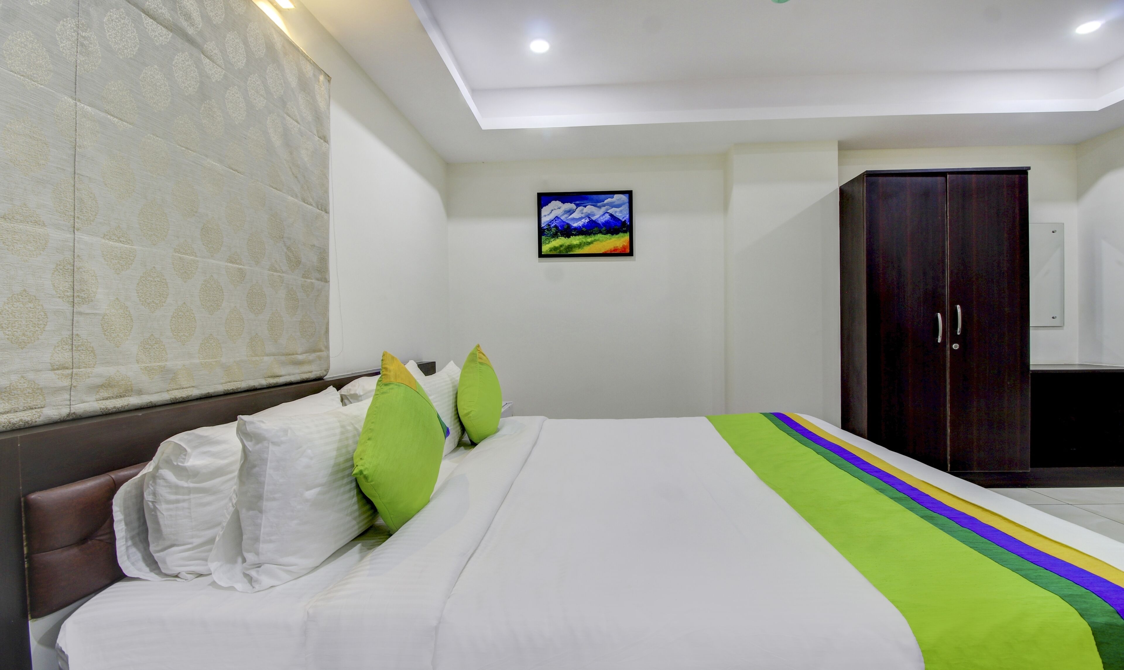 A neatly made bed with white bedding and green pillows, a dark wooden wardrobe, and a framed picture on the wall.