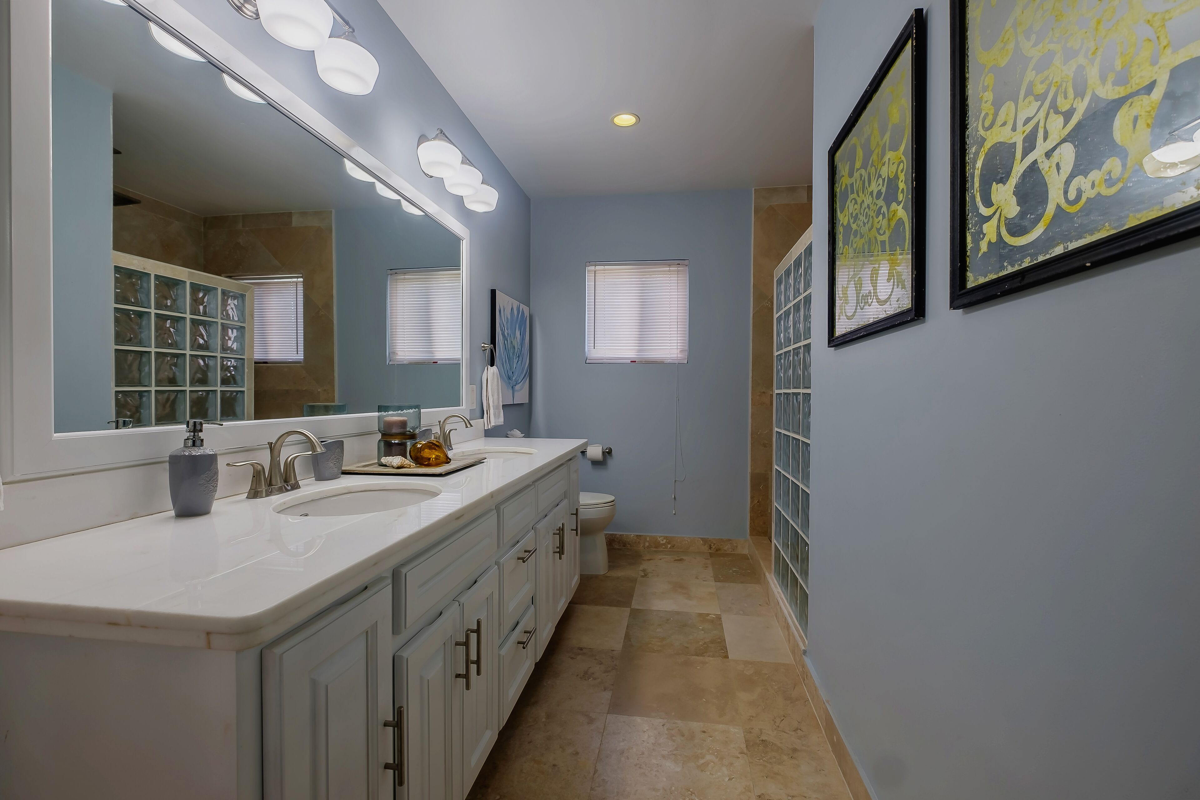 Une salle de bain avec un grand miroir, deux lavabos et des toilettes. Les murs sont ornés d’œuvres d’art encadrées et il y a une fenêtre avec des stores.