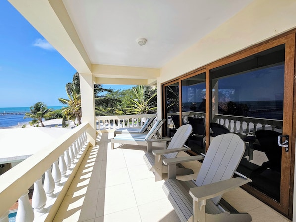 Uma varanda com cadeiras e mesa brancas, com vista para uma praia com palmeiras e o oceano.