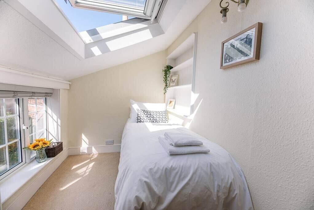 A bedroom with a skylight, a bed with white bedding, a window with flowers, and a wall-mounted picture.