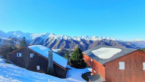 A wooden cabin with a snow-covered roof, surrounded by snow-covered mountains and clear blue sky.