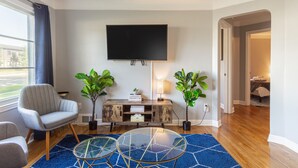 Un salon moderne avec un téléviseur à écran plat, deux plantes en pot, un fauteuil gris, une table basse en verre et un tapis bleu.