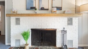 Une cheminée en briques blanches surmontée d’une cheminée en bois, avec trois œuvres d’art encadrées au-dessus, et une plante en pot sur la gauche.