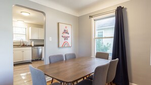 Un coin repas avec une table en bois et des chaises grises, une cuisine avec des meubles blancs et une fenêtre ornée de rideaux foncés.