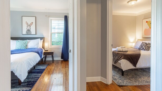 Une chambre avec deux lits, une fenêtre avec des rideaux bleus, une table de chevet avec une lampe et un tableau encadré au mur.