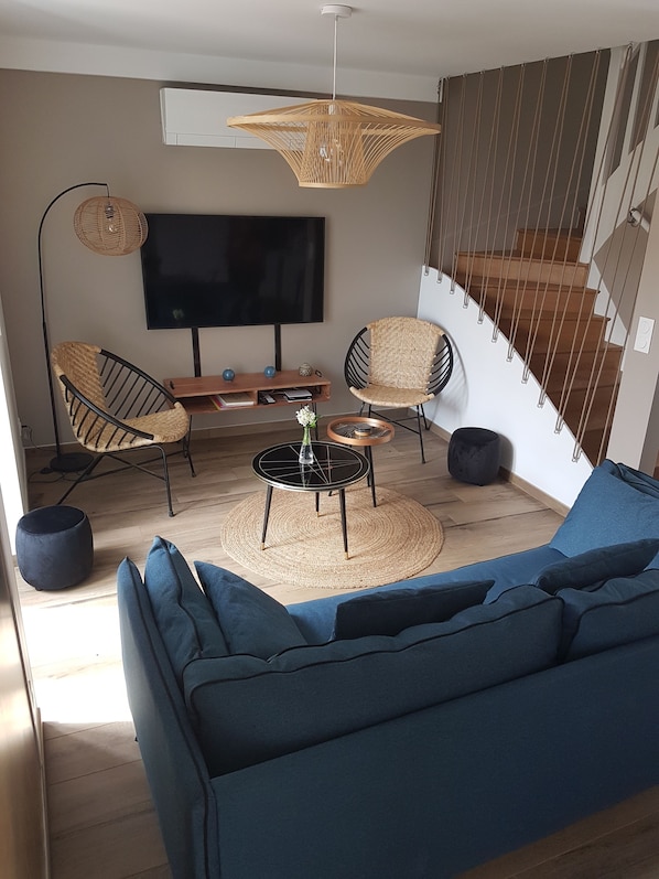 Een moderne woonkamer met een blauwe bank, rieten stoelen, een ronde salontafel en een grote flatscreen televisie.