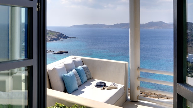 Un balcone con un divano bianco, cuscini e un cappello, con vista sulla costa, mare azzurro e colline lontane.