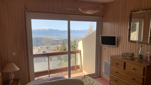 A bedroom with a bed, a dresser, a TV, and a sliding glass door leading to a balcony with a mountain view. - Appartement à 2 Minutes à Pied des Commerces et Télécabines Avec Superbe vue (Font-Romeu-Odeillo-Via)