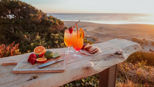 A wooden table with a cutting board, fruit, and two glasses of orange juice with a straw and a strawberry garnish, set against a beach backdrop. - Ocean Front with Beautiful Views! Miles of Sandy Beach! (Seal Rock)