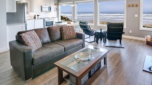A modern living room with a gray sofa, a glass coffee table, and a view of the ocean through large windows. - Ocean Front with Beautiful Views! Miles of Sandy Beach! (Seal Rock)