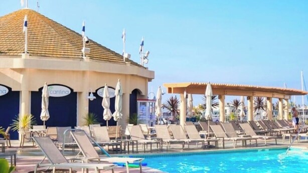 A swimming pool with lounge chairs and umbrellas, a covered area, and a building with a tiled roof.