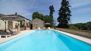 A rectangular swimming pool with clear blue water, surrounded by a stone wall. There are white lounge chairs and umbrellas on the poolside.