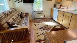 A compact living space with a sofa, a dining table, a kitchen area with wooden cabinets, a flat-screen TV mounted on the wall, and a patterned rug.