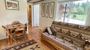 A cozy living room with a wooden sofa, a dining table with chairs, and a large window offering a view of the outdoors.