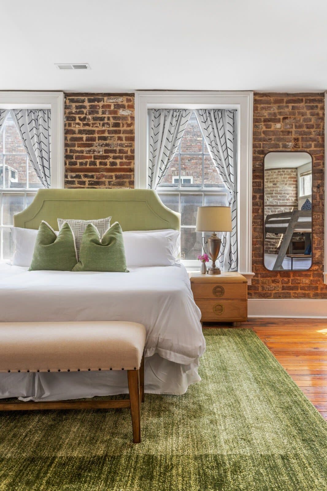 A bedroom with a brick wall, a bed with a green headboard, a bench, a nightstand with a lamp, and a mirror on the wall.