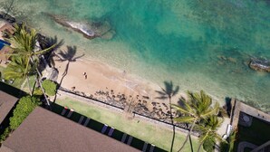 Luchtbeeld van een strand met helder turquoise water, palmbomen en een rotsachtige kustlijn.