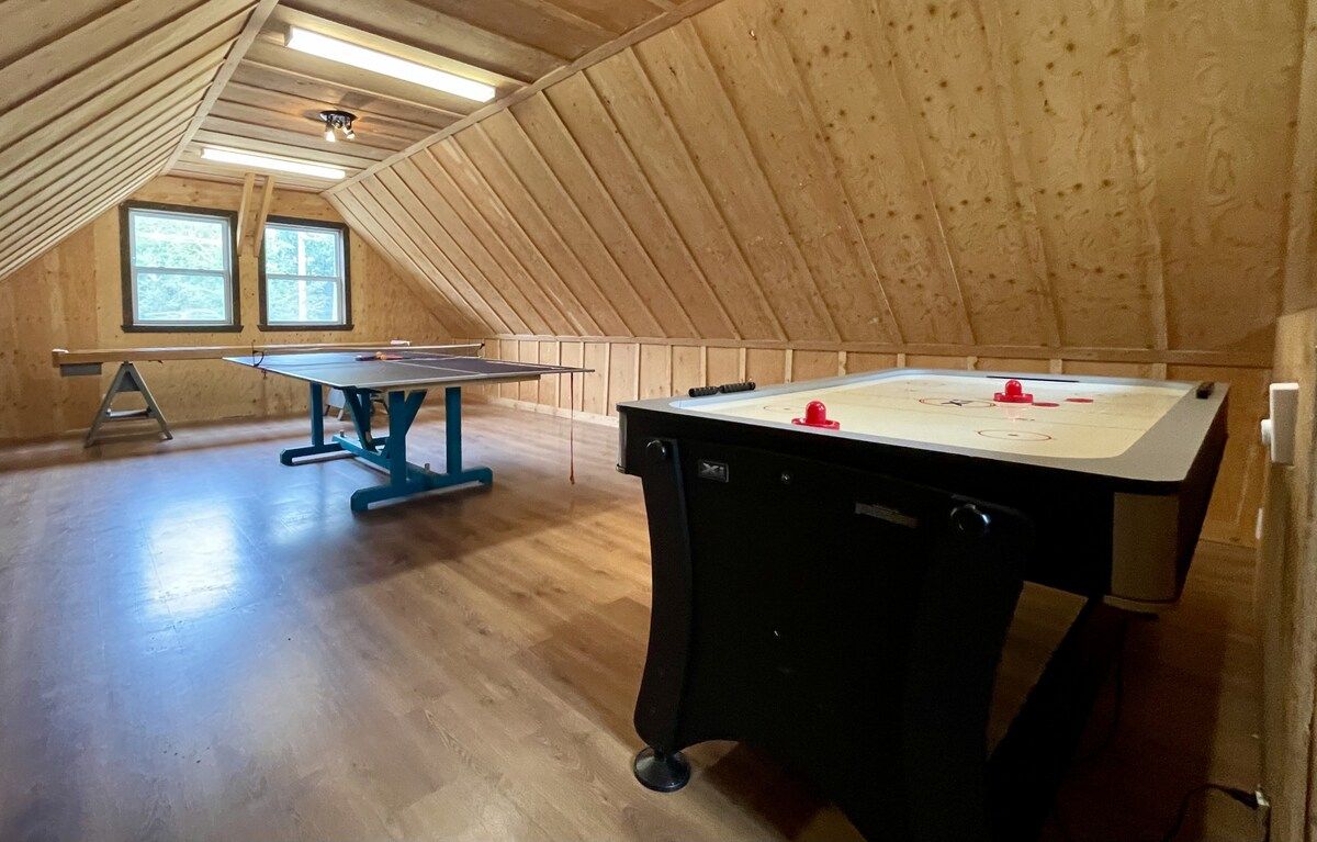 An attic room with wooden walls and a sloped ceiling, featuring a ping pong table and a foosball table.