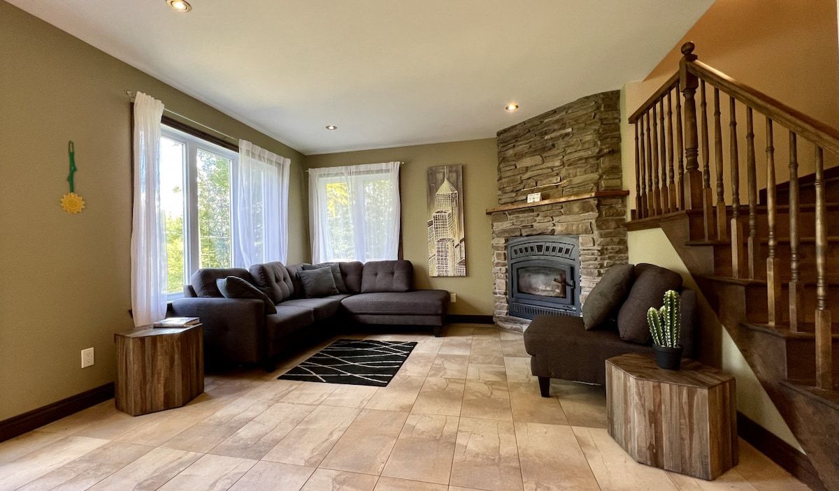 A living room with a sectional sofa, a fireplace, a staircase, and a view of trees through the windows.