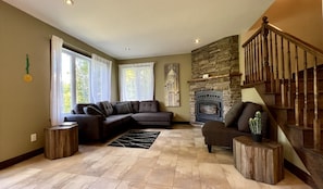 A living room with a sectional sofa, a fireplace, a staircase, and a view of trees through the windows.