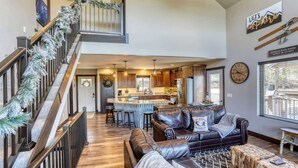 A spacious living area with a staircase, a kitchen in the background, and a view of the outside. - Annie Creek Lodge – Hot tub, game loft, fireplaces & stunning views! (Lead)