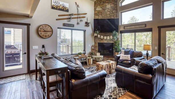 A spacious living room with a stone fireplace, leather sofas, a dining area, and large windows offering views of the outdoors. - Annie Creek Lodge – Hot tub, game loft, fireplaces & stunning views! (Lead)
