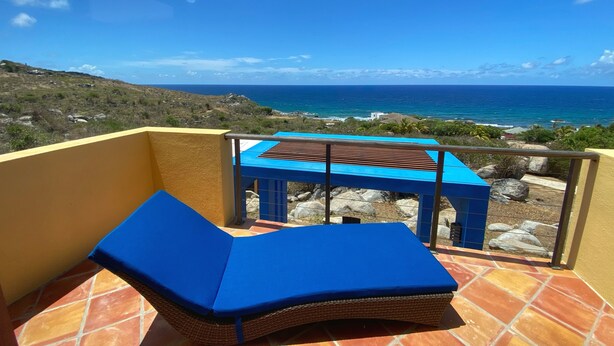 Een balkon met een blauwe loungestoel met kussens en een klein tafeltje, met uitzicht op een kustlandschap met rotsachtig terrein en de oceaan.