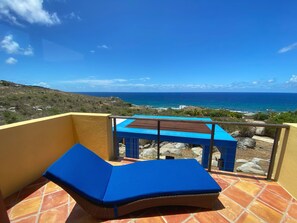 Un balcon avec un fauteuil de relaxation bleu doté de coussins et une petite table, offrant une vue sur un paysage côtier caractérisé par un terrain rocheux et l’océan.