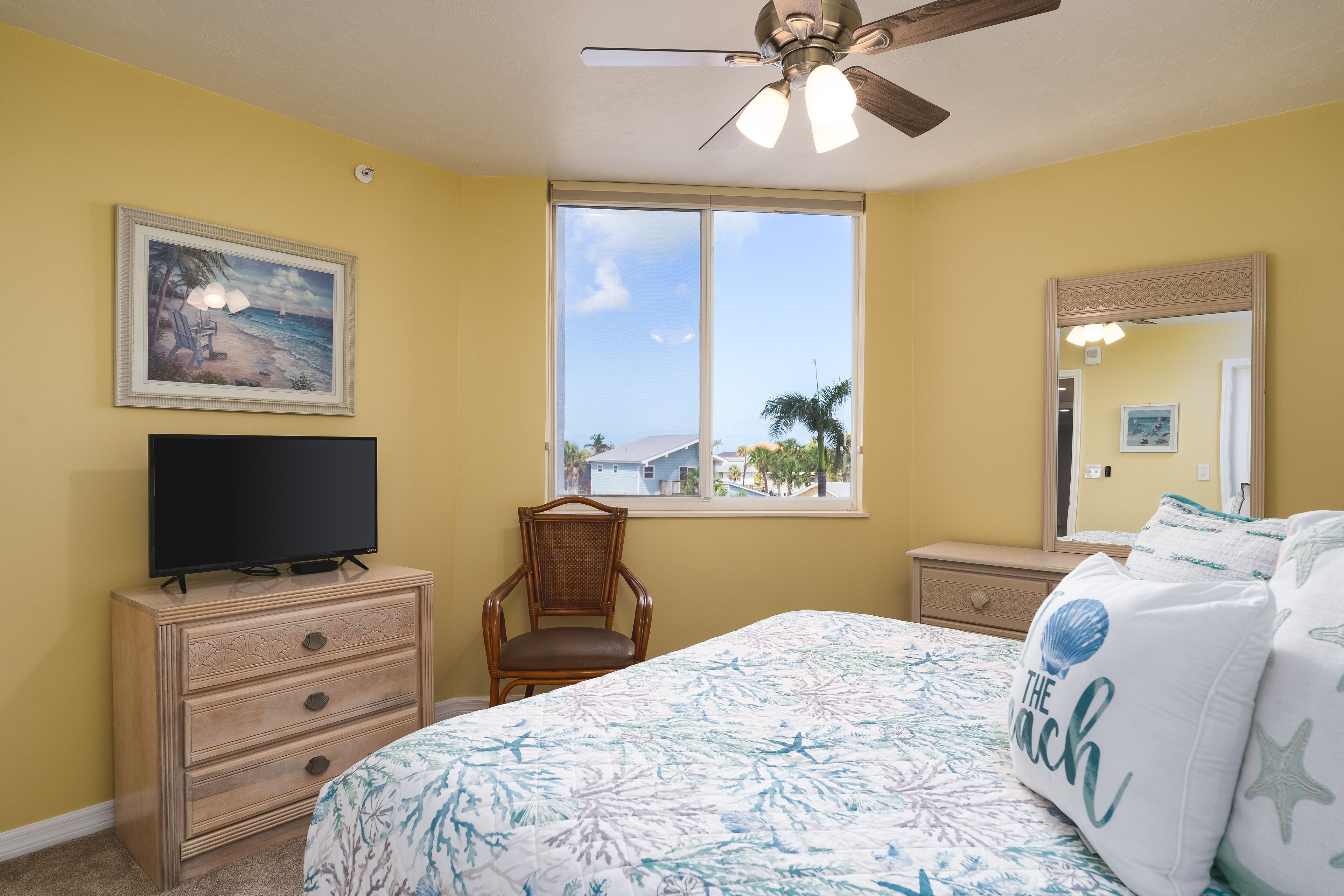 A bedroom with a bed, a television on a dresser, a chair, a window with a view, and a ceiling fan.