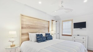 Un dormitorio con cabecera de madera, cama con ropa de cama azul y blanca a rayas, mesita de noche con lámpara, televisor en la pared y ventana con persianas.