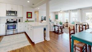 A spacious kitchen with white cabinetry, a microwave, an oven, and a large island. There is a dining area with a table and chairs, and a living room with a sofa and a coffee table. - Heated Pool Access, Tybee Beach Bungalow (Tybee Island)
