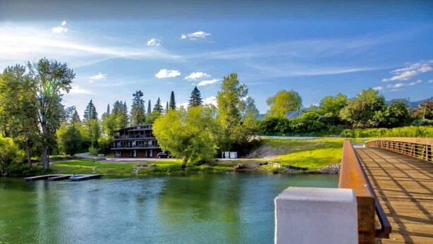 A riverside view with a wooden bridge, a building with a sloped roof, and a grassy area with trees. - On The River | Hot Tub | Fire Pit | Upgraded AC! (Whitefish)