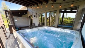 A hot tub on an outdoor deck with chairs and a table, surrounded by windows. - On The River | Hot Tub | Fire Pit | Upgraded AC! (Whitefish)