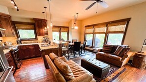 A spacious living area with a sofa, ottoman, and dining table, connected to a kitchen and dining area with large windows. - On The River | Hot Tub | Fire Pit | Upgraded AC! (Whitefish)