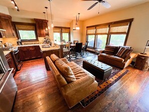 A spacious living area with a sofa, ottoman, and dining table, connected to a kitchen and dining area with large windows. - On The River | Hot Tub | Fire Pit | New AC 2024! (Whitefish)