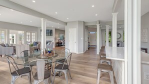 Een ruime woonkamer met een glazen eettafel, moderne stoelen en een keuken met een wit aanrecht en krukken.