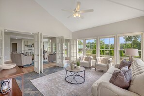 Un salon spacieux doté d’une grande fenêtre, d’un ventilateur de plafond et d’un coin repas avec une table et des chaises.