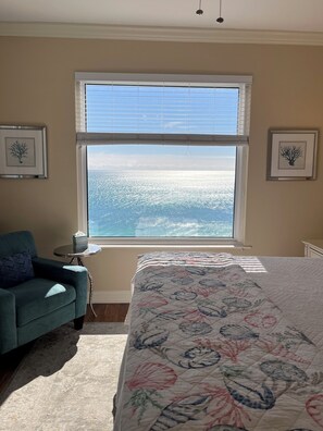 Une chambre avec un grand lit, une vue sur l’océan par la fenêtre, un fauteuil et deux tableaux encadrés au mur.