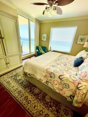 A bedroom with a large bed, a view of the ocean through a sliding door, a ceiling fan, and a small side table with a lamp.