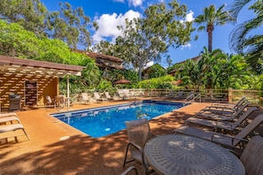 Relax by the serene pool surrounded by lush greenery and palm trees, with lounge chairs and a table for a peaceful retreat.
